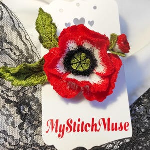 May include: A red, white, and black embroidered poppy flower with green leaves and a bud, attached to a white tag. The tag has the text "MyStitchMuse" in red script. The flower is set against a white background with black lace.