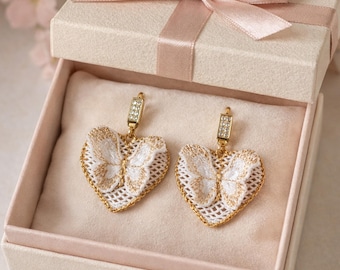 Puede incluir: Pendientes en forma de corazón con diseño de mariposa, presentados en una caja de regalo. Los pendientes presentan bordados blancos con detalles dorados y un borde de cadena dorada. La caja es de color crema con una cinta rosa.