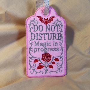 Op de afbeelding: Roze vilten label met de woorden "DO NOT DISTURB Magic in progress" in zilveren glitter. Een rode vleermuis en roos zijn ook afgebeeld. Het label is afgewerkt met zilveren glitter en heeft een zilveren lint om op te hangen.
