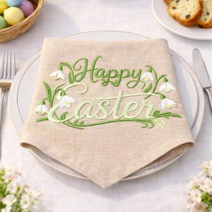 Può includere: Un tovagliolo di lino beige con le parole "Happy Easter" ricamate in verde, circondate da foglie verdi e fiori bianchi. Il tovagliolo è piegato su un piatto bianco, pronto per un pasto festivo.