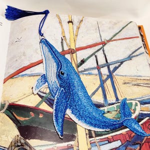 Könnte beinhalten: Ein Lesezeichen in Form eines Blauwals mit detaillierter Stickerei in Blau- und Weißtönen. Der Wal ist mit einem silbernen Metallring an einer dunkelblauen Seidenquaste befestigt und liegt auf einer Illustration von Booten am Sandstrand.