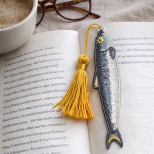 Può includere: Segnalibro a forma di pesce ricamato con nappina gialla, su un libro aperto. Il segnalibro è grigio e bianco con dettagli gialli. Una tazza di caffè e occhiali sono sullo sfondo.