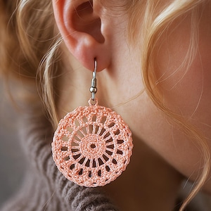May include: A close-up of a peach-colored, crocheted earring. The circular earring features an openwork design with a central pattern and a metal hook. The earring is hanging from a person's ear, with blonde hair and a brown sweater in the background.