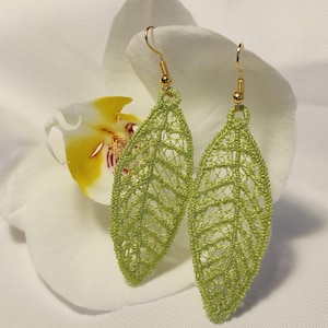 May include: A pair of light green leaf-shaped earrings with gold-colored hooks. The earrings are made of a delicate, open-weave material, and are set against a white background with a white and yellow flower.