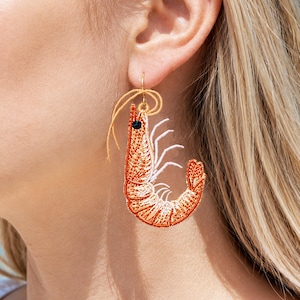 May include: A single shrimp earring with orange and white embroidery, a black bead eye, and gold-colored wire. The earring is hanging from a gold-colored hook. The earring is worn on a person's ear.