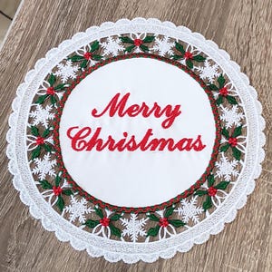 May include: A white, round Christmas doily with red "Merry Christmas" script. The doily features a red and green holly and snowflake design, with a white lace edge. The doily is placed on a wooden surface.