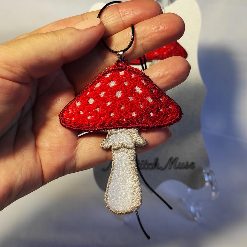 Op de afbeelding: Een rode paddestoel hanger met witte stippen en een cr&egrave;mekleurige steel. De hanger is geborduurd en heeft een zwart koord. Een grillig accessoire.