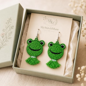 May include: A pair of green embroidered frog earrings with matching green tassels, displayed on a MyStitchMuse branded card inside a sage green gift box. The frog faces feature black stitched eyes and a curved mouth, hanging from silver-toned hooks.