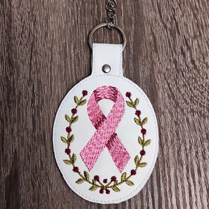 以下が含まれることがあります： 中央にピンクのリボンが刺繍され、緑の葉と赤い実のリースで囲まれた白い楕円形のキーホルダー。 キーホルダーには、白い革のような素材に取り付けられたシルバーの金属リングが付いています。