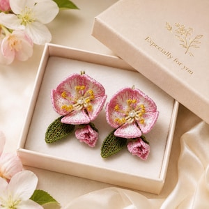 May include: A pair of handcrafted floral earrings in a gift box. The earrings feature pink and white embroidered flower designs with yellow accents and a central pearl. The gift box is light pink with the text "Especially for you".