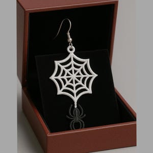 May include: A white spiderweb earring with a black spider charm, displayed in a brown jewelry box with a black interior. The earring has a silver hook.