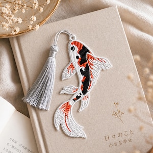 Könnte beinhalten: Bestickter Koi-Karpfen-Lesezeichen mit silberner Quaste. Das Fischdesign weist weiße, schwarze und orangefarbene Details auf. Das Lesezeichen liegt auf einem geschlossenen Buch mit japanischem Text und einem kleinen Blumendesign.