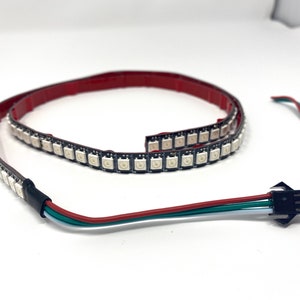 May include: Flexible LED strip with multiple small, square lights. The strip is black with a red adhesive backing. A separate connector with red, green, and blue wires is also visible. The lights are designed for various lighting projects.