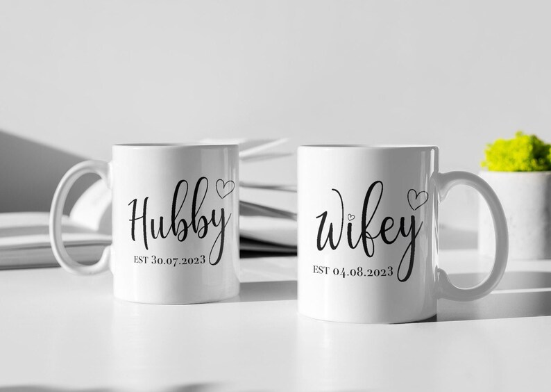 May include: Two white ceramic mugs with black script that reads "Hubby" and "Wifey", each with a heart symbol. The "Hubby" mug has "EST 30.07.2023" below the word. The "Wifey" mug has "EST 04.08.2023" below the word.