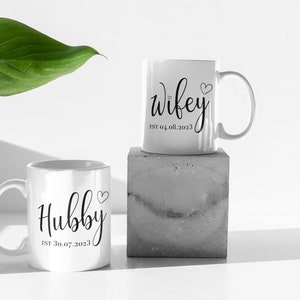 May include: Two white ceramic mugs with black script. One mug reads "Hubby" with a heart and "EST 30.07.2023". The other mug reads "Wifey" with a heart and "EST 04.08.2023". One mug is on a concrete block.