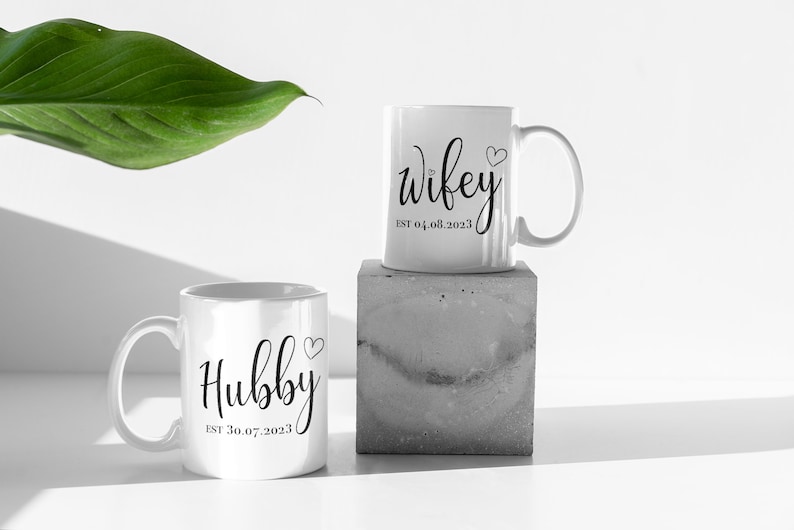 May include: Two white ceramic mugs with black script. One mug reads "Hubby" with a heart and "EST 30.07.2023". The other mug reads "Wifey" with a heart and "EST 04.08.2023". One mug is on a concrete block.