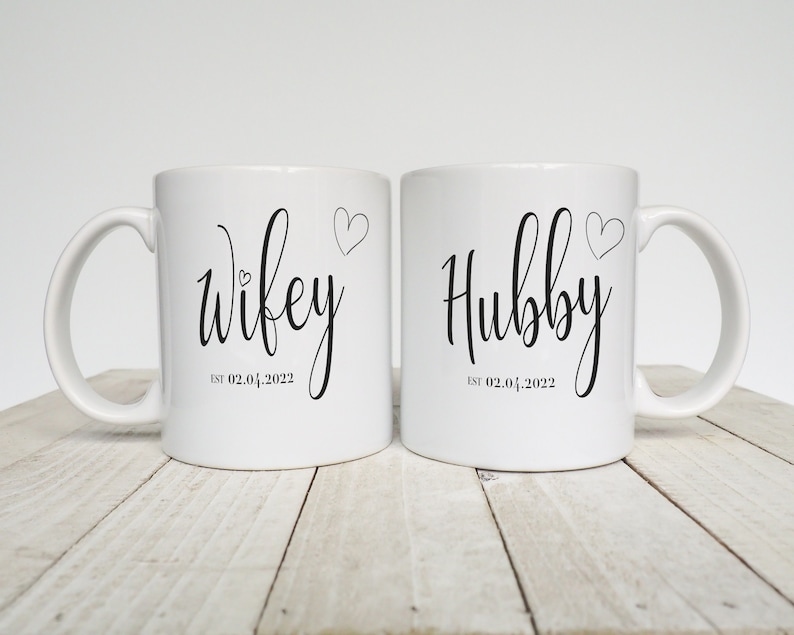 May include: Two white ceramic mugs with black script that reads "Wifey" and "Hubby". Each mug has a heart graphic and the date "EST 02.04.2022". The mugs are on a light wood surface.