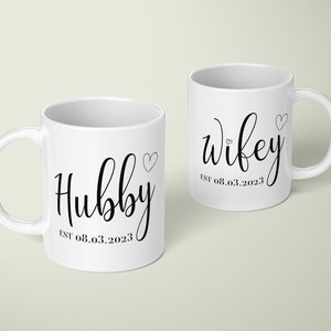 May include: Two white ceramic mugs with black script. One mug says "Hubby" with a heart and "EST 08.03.2023". The other says "Wifey" with a heart and "EST 08.03.2023".