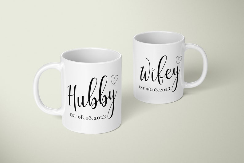 May include: Two white ceramic mugs with black script. One mug says "Hubby" with a heart and "EST 08.03.2023". The other says "Wifey" with a heart and "EST 08.03.2023".