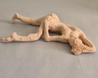 May include: A light-colored clay sculpture depicting a human figure in a prone position. The figure is lying on its stomach with arms and legs extended, and the head is turned to the side. The sculpture has a rough, textured surface.