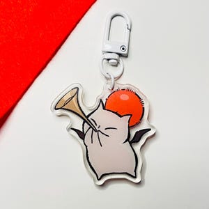 May include: A white acrylic keychain featuring a cartoon white cat with black wings playing a golden trumpet. The cat has a red halo and is set against a white background.