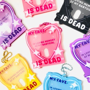 Könnte beinhalten: Ein Set aus sechs bunten herzförmigen Schlüsselanhängern mit dem Text "My Fave" und "Is Dead" in weißen Buchstaben. Jeder Schlüsselanhänger hat ein Sternendesign und ein transparentes Fenster für ein Foto.