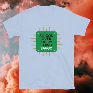Op de afbeelding: Lichtblauw T-shirt met een groene vierkante afbeelding met de witte tekst "SILICON OVER EVERYTHING $AVGO". De afbeelding is omgeven door gouden lijnen, die lijken op een printplaat. Een casual kledingstuk.
