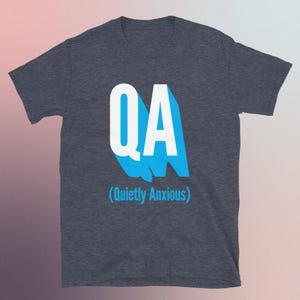 May include: Dark grey t-shirt featuring the white letters "QA" with a light blue shadow. The text "(Quietly Anxious)" is below the letters in a smaller font. The t-shirt is made of a soft material and is suitable for casual wear.