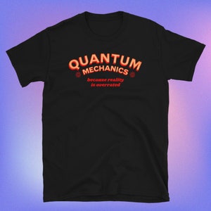 以下が含まれることがあります： 「QUANTUM MECHANICS because reality is overrated」と書いてある、赤とオレンジのグラフィックがプリントされた黒いTシャツです。