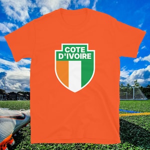 May include: An orange t-shirt featuring a shield design with the text "COTE D'IVOIRE" in green and white. The shield incorporates the colors orange, white, and green, representing the flag of Ivory Coast. The shirt is displayed on a soccer field.