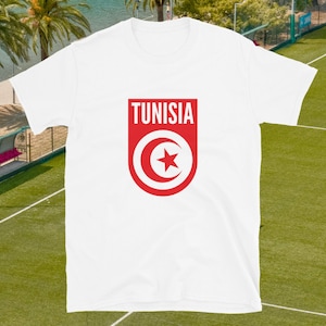 May include: White t-shirt with a red shield design. The word "TUNISIA" is at the top of the shield, with a red crescent moon and star emblem below. The shirt is short-sleeved and made of a soft material.