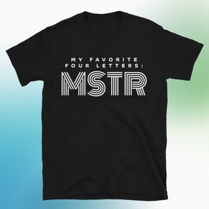 May include: Black t-shirt with white text that reads "MY FAVORITE FOUR LETTERS: MSTR". The letters MSTR are designed with a striped pattern. The shirt is a crew neck style.