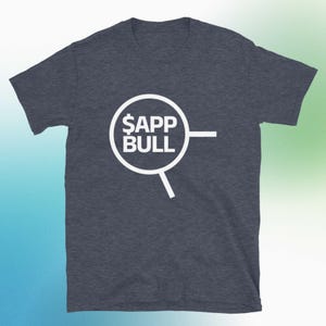 以下が含まれることがあります： 白のグラフィックが入った濃いヘザーグレーの半袖Tシャツ。グラフィックは、虫眼鏡のデザインの白い円の中に「$APP BULL」というテキストを表示しています。Tシャツは柔らかい素材で作られています。