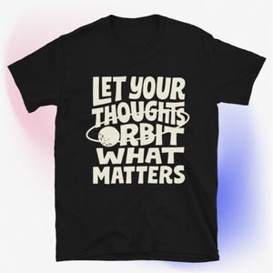 May include: Black t-shirt with the white text "LET YOUR THOUGHTS ORBIT WHAT MATTERS." The design includes a planet graphic. The shirt is a crew neck and has short sleeves.