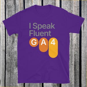 May include: A purple t-shirt with the text "I Speak Fluent GA4" in gray, orange, and yellow. The shirt is made of a soft material and has a crew neck.