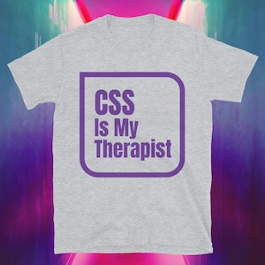 Op de afbeelding: Lichtgrijs t-shirt met de tekst "CSS Is My Therapist" in een paarse, afgeronde rechthoek. Het shirt heeft een ronde hals en korte mouwen.