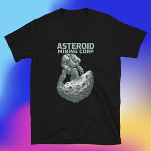 Op de afbeelding: Zwart t-shirt met de tekst "ASTEROID MINING CORP" en een gedetailleerde illustratie van een astronaut op een maanachtig rotsblok. De astronaut draagt een ruimtepak.