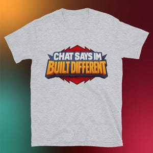 Peut inclure: T-shirt gris clair avec l'inscription "CHAT SAYS IM BUILT DIFFERENT" dans un graphisme bleu, orange et rouge. Le t-shirt est en matière douce et a des manches courtes.
