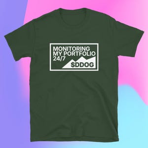 以下が含まれることがあります： 白い長方形のグラフィックが付いた濃い緑色のTシャツ。グラフィックには、山のようなグラフとテキスト「$DDOG」の上に「MONITORING MY PORTFOLIO 24/7」と書かれています。シャツは柔らかい素材で作られています。