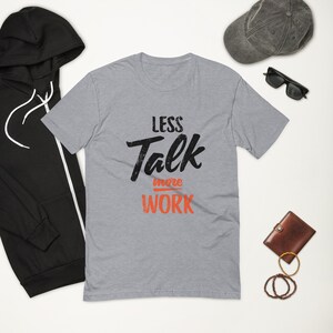 Puede incluir: Una camiseta gris jaspeada con un gráfico negro que dice "Less Talk More Work" en una fuente de estilo vintage. La palabra "more" está en naranja.