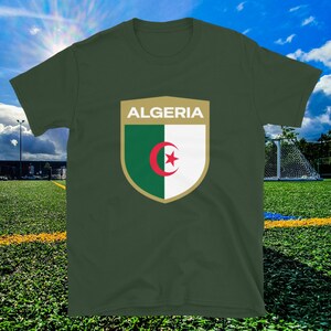 May include: An olive green t-shirt with a shield design displaying the word "ALGERIA". The shield features the Algerian flag: green and white sections with a red crescent and star. The shirt is on a green field.