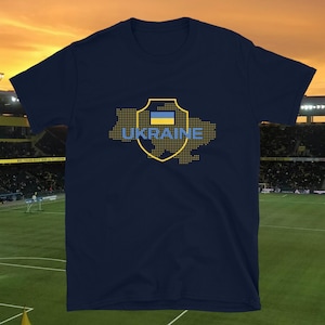 Puede incluir: Camiseta azul marino con un gráfico amarillo y azul de la bandera ucraniana y la palabra "UKRAINE". El gráfico tiene la forma del contorno del país. La camiseta está hecha de un material suave.