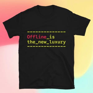 Op de afbeelding: Zwart T-shirt met de tekst "Offline_is the_new_luxury" in geel en roze. De tekst is gecentreerd en heeft een stippellijn erboven en eronder. Het shirt is gemaakt van een zacht materiaal.