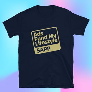 May include: Navy blue t-shirt with a gold-colored square graphic. The graphic features the text "Ads Fund My Lifestyle" above "$APP". The t-shirt is made of a soft material and is suitable for casual wear.