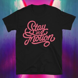 Puede incluir: Camiseta negra con el texto rosa "Stay in Motion" en una fuente cursiva. El diseño está centrado en la parte delantera. La camiseta tiene mangas cortas y cuello redondo.