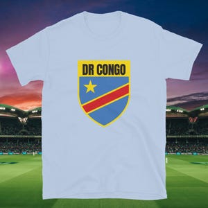Pode incluir: T-shirt azul claro com um design de escudo com o texto "DR CONGO" em amarelo. O escudo tem um fundo azul com uma estrela amarela e uma faixa vermelha. A camisa é feita de um material macio.