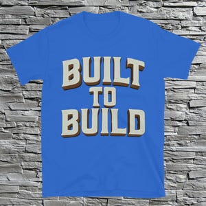May include: A blue t-shirt with the words "BUILT TO BUILD" in a bold, vintage-style font. The text is in white with a brown shadow, creating a 3D effect. The shirt is made of a soft material.