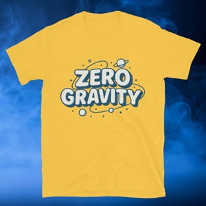 May include: Yellow t-shirt with the words "ZERO GRAVITY" in a white and blue bubble font. The design includes a planet and orbiting dots, creating a space-themed graphic.