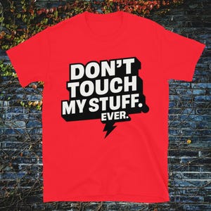 May include: A red t-shirt with the text "DON'T TOUCH MY STUFF. EVER." in bold white letters with a black outline. A lightning bolt graphic is below the text. The shirt is displayed against a brick wall.
