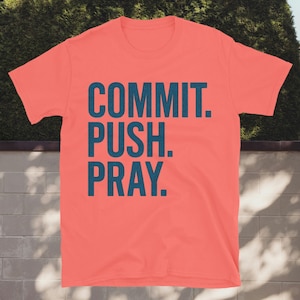 Może przedstawiać: Koralowa koszulka z napisem "COMMIT. PUSH. PRAY." w ciemnoniebieskim kolorze. Tekst jest ułożony pionowo, każde słowo w osobnej linii. Koszulka jest prezentowana na tle zieleni i ceglanego muru.